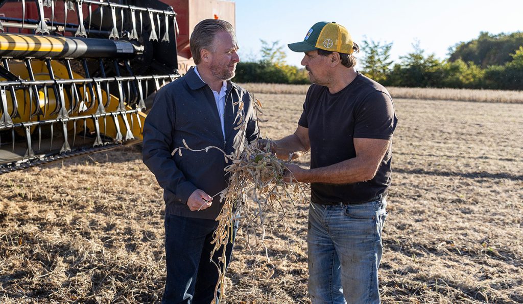 Jason Brass meeting a local farmer on the farm. They are standing in a field with farm equipment in the background.
