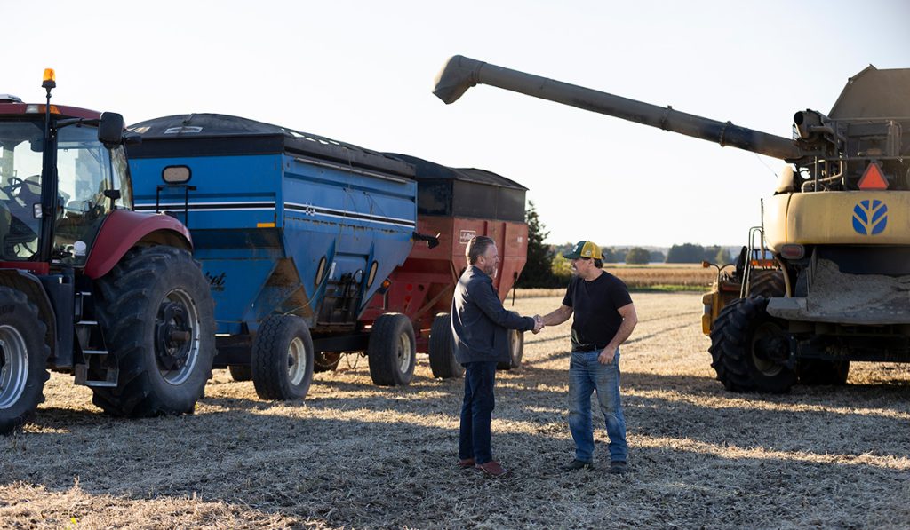Jason Brass meeting a local farmer on the farm. They are standing in a field with farm equipment in the background.